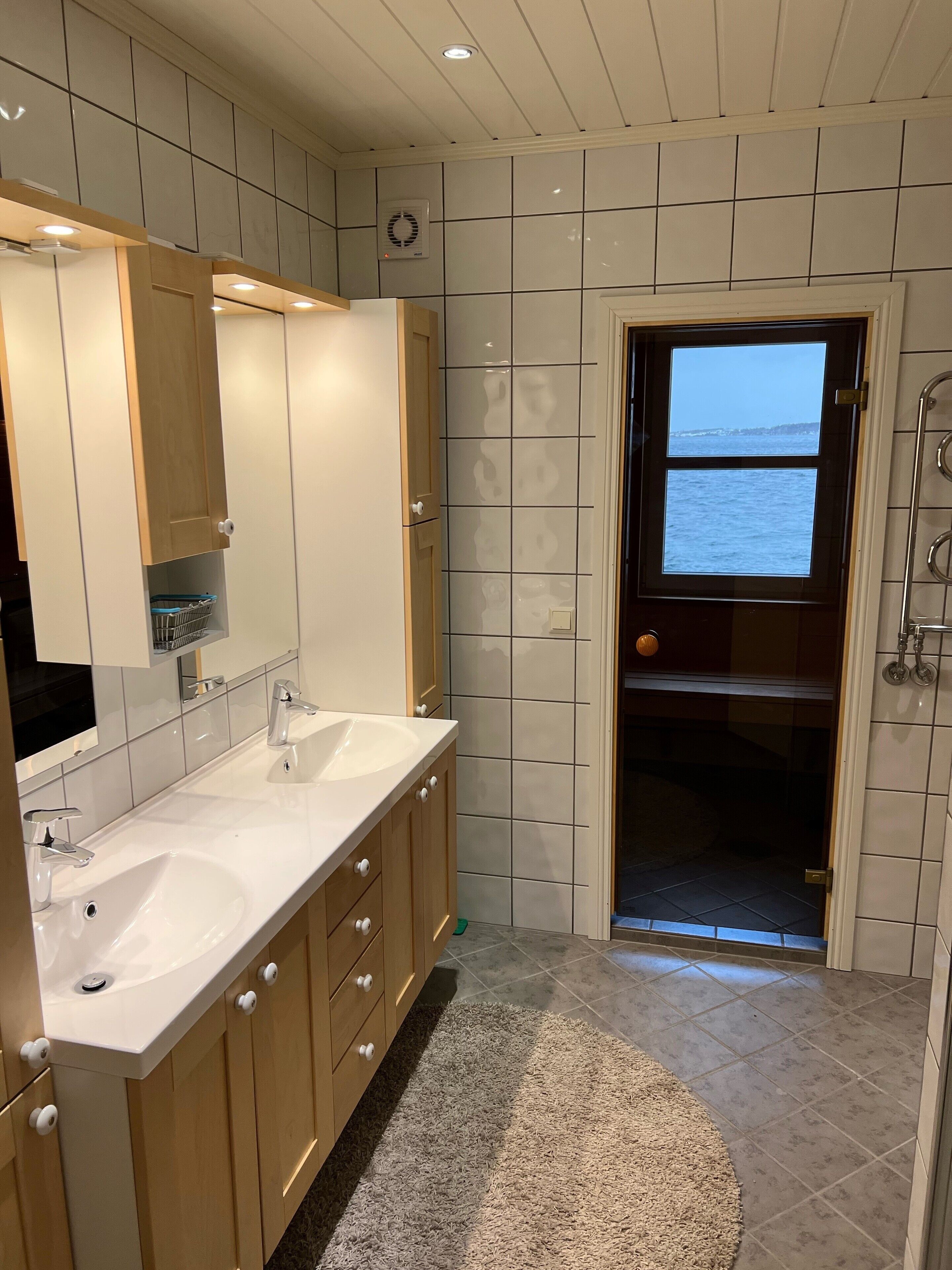 A modern bathroom with two sinks, wooden cabinets, a large mirror, and a view of the sea through a door.