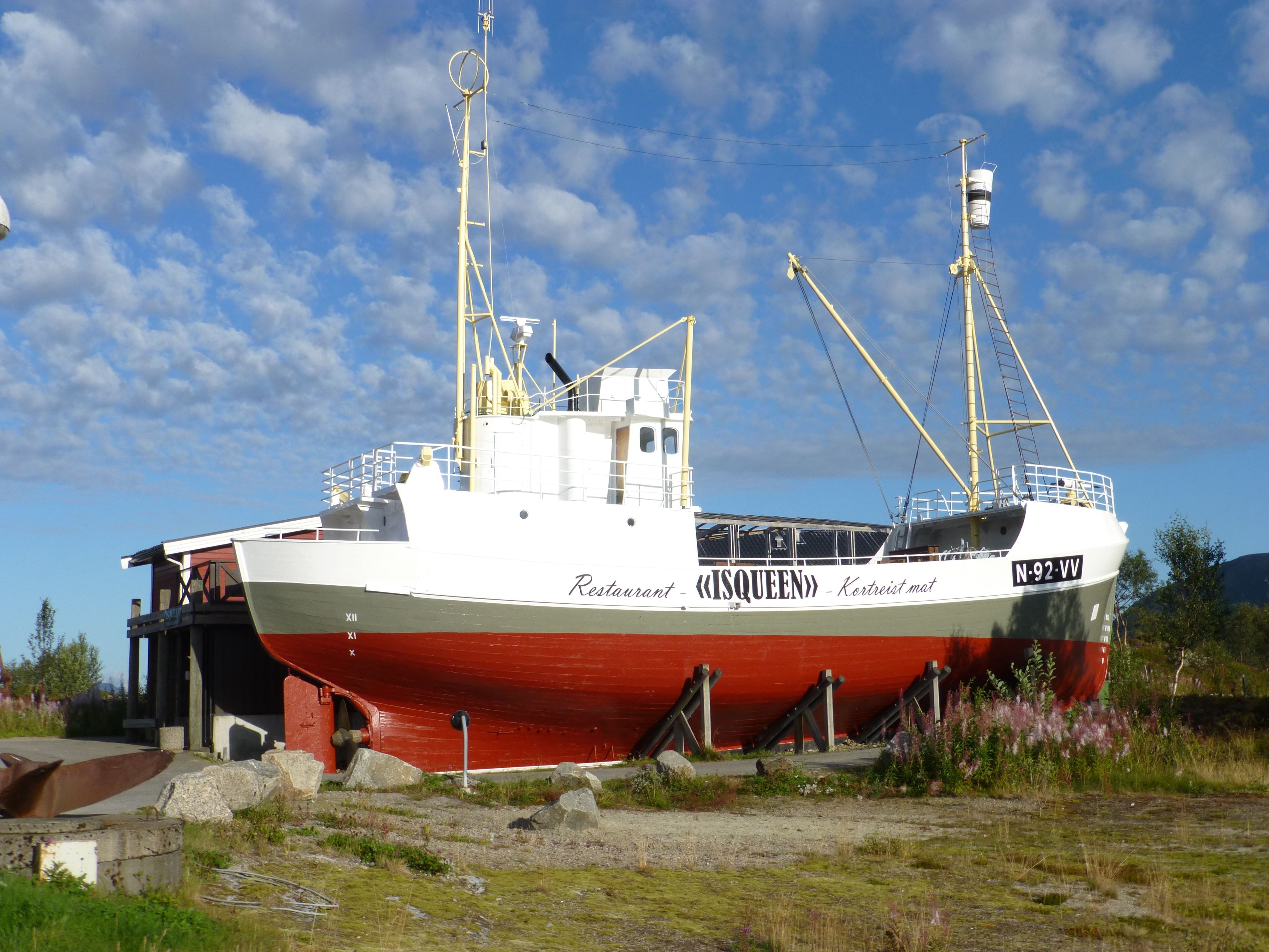 A large, stationary ship with the name "Isqueen" and the number "N-92-VV" on its side, docked in a grassy area with a clear sky in the background.