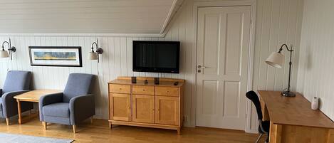 Una habitación con un armario de madera, un televisor, dos sillas grises, un escritorio de madera y una lámpara.