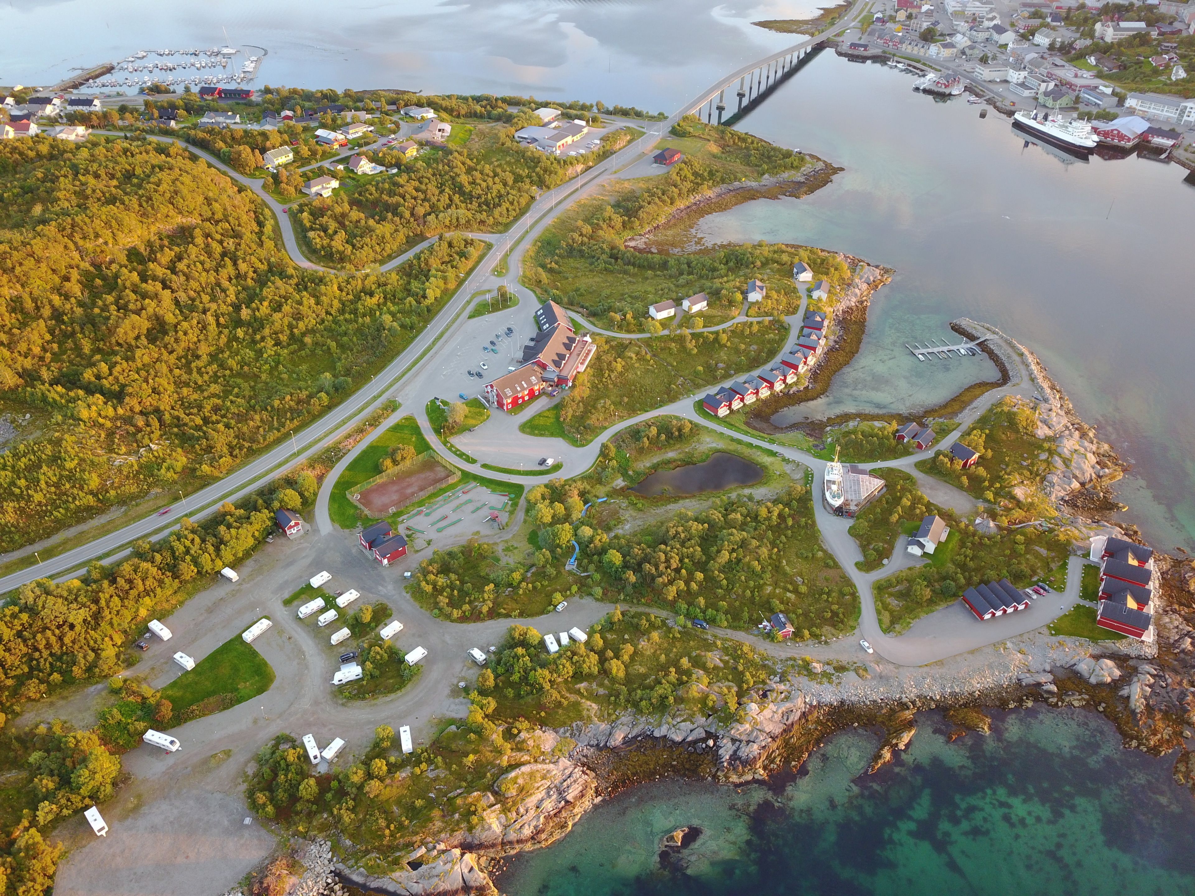 Vue aérienne d’une zone côtière comprenant un port de plaisance, un réseau routier et plusieurs bâtiments, dont une grande structure rouge et des maisons plus petites.