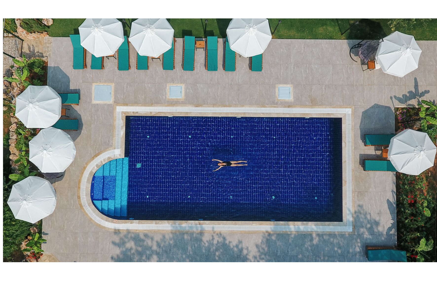 Vista aérea de una piscina rectangular con una persona nadando, rodeada de sillas de descanso y sombrillas.