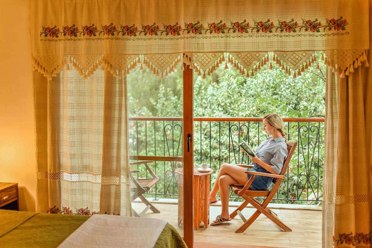 A room with a balcony, a bed, a chair, and a table with a cup.