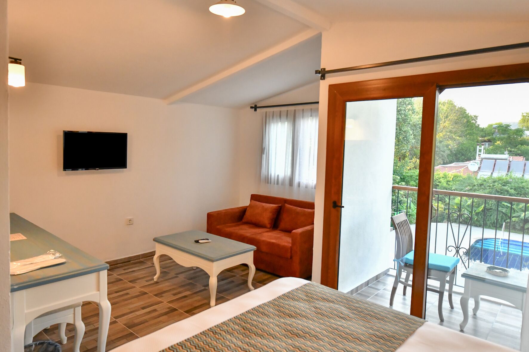 Une chambre avec un lit, une petite table, un canapé, une télévision et un balcon donnant sur une piscine.