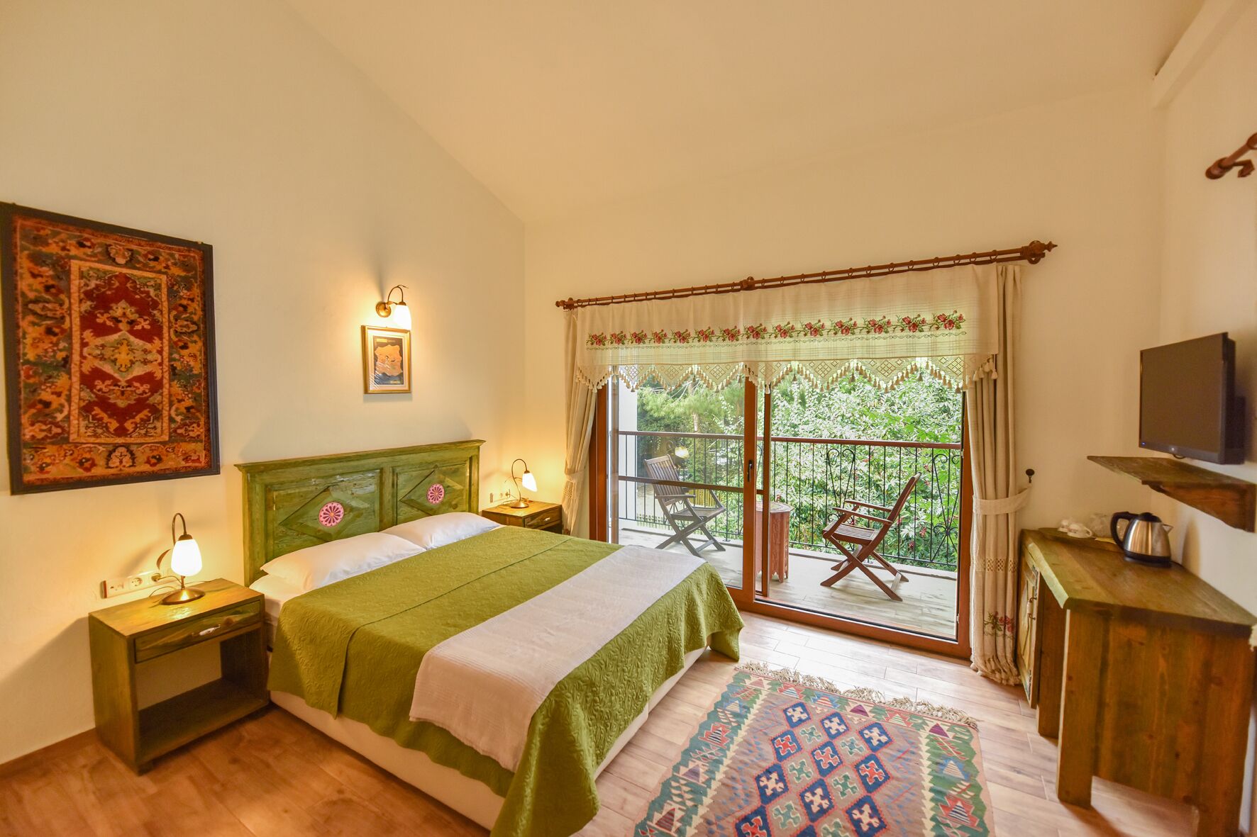 A bedroom with a bed, bedside lamps, a TV, a balcony with chairs, and a view of greenery.