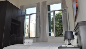 A hotel room with a bed, a nightstand with a lamp, a wardrobe, and two windows with curtains. - TLV Urban Life Style (Tel Aviv)