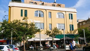 A yellow building with multiple windows and a flat roof. There are outdoor seating areas with umbrellas and people walking around. - TLV Urban Life Style (Tel Aviv)