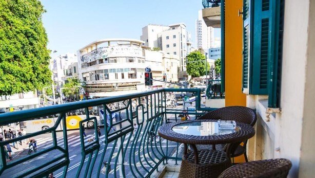 A balcony with wicker furniture and a glass table, overlooking a busy street with buildings and trees. - TLV Urban Life Style (Tel Aviv)