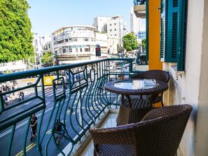 Un balcon agrémenté de meubles en osier et d’une table en verre, donnant sur une rue animée bordée d’immeubles et d’arbres.