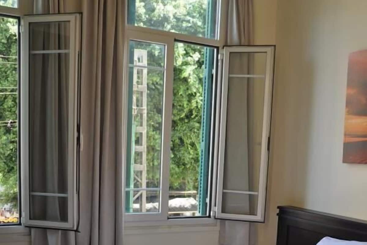 A neatly made bed with white linens and towels, a window with curtains, and a view of greenery outside.