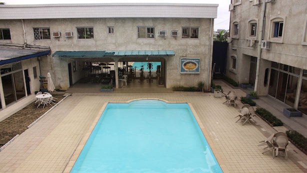 A swimming pool with a tiled deck, a covered seating area, and a building with multiple windows and air conditioning units. - Jet Hôtel (Douala)