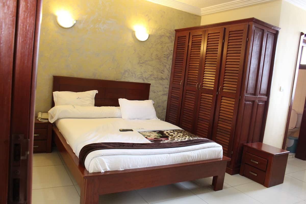 A neatly made bed with white linens and a remote control, a wooden nightstand with a phone, and a large wooden wardrobe with louvered doors.