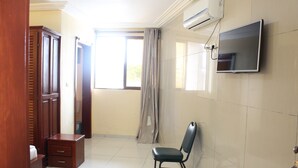 A hotel room with a wooden wardrobe, a small bedside table, a chair, a flat-screen TV mounted on the wall, and a window with curtains. - Hotel Joana (Douala)