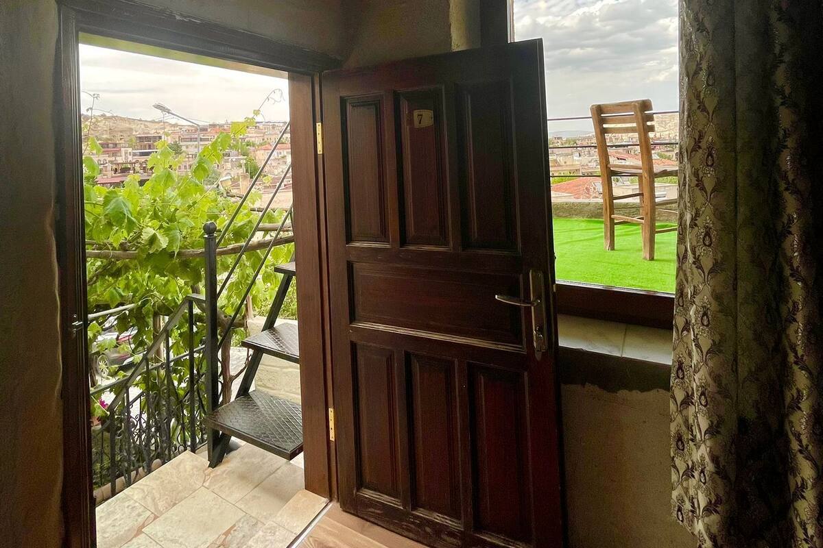 Una habitación con puerta de madera, ventana con vistas a un césped verde y edificios, y baranda metálica en el exterior.
