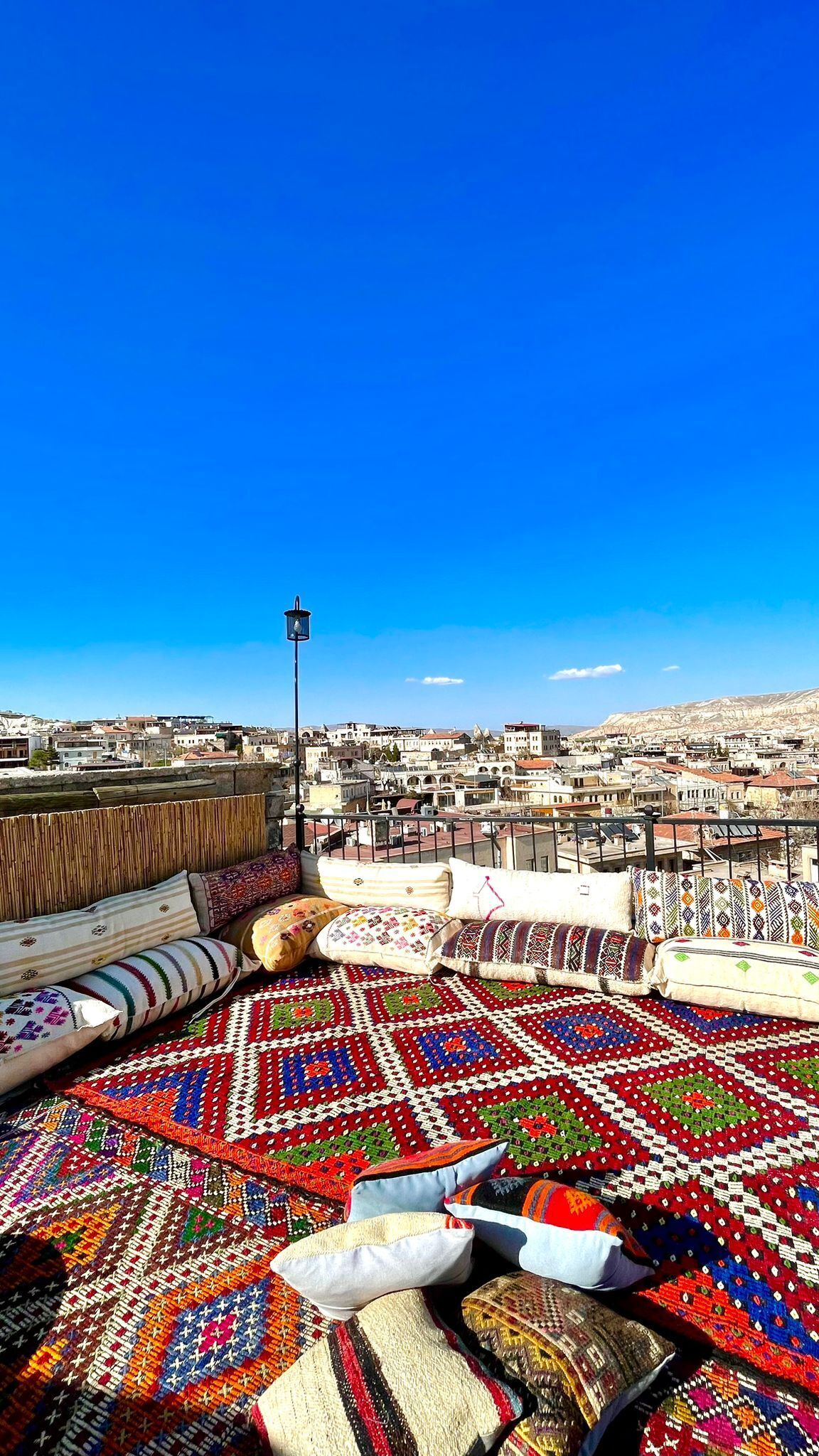 A rooftop terrace with a view of a cityscape, featuring a patterned rug and various cushions.