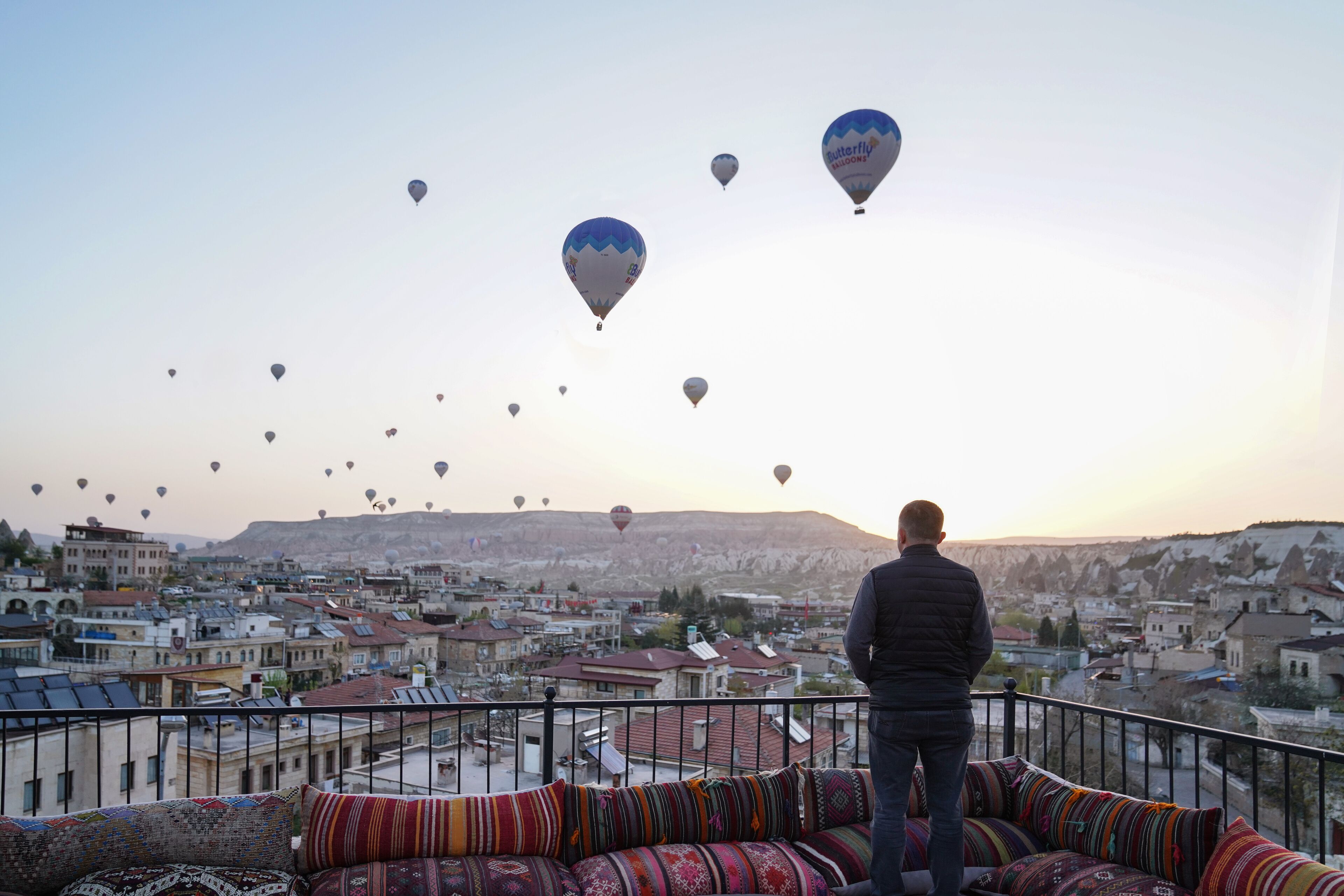 Un balcón con cojines de colores con vistas a un paisaje urbano y globos aerostáticos en el cielo.