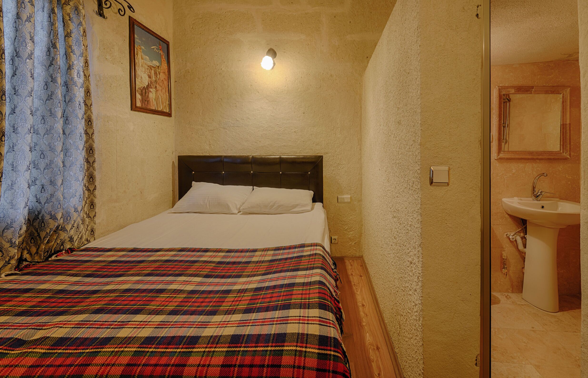 Une chambre à coucher avec un lit, une table de chevet, une lampe fixée au mur, et une salle de bain avec un lavabo et un miroir.