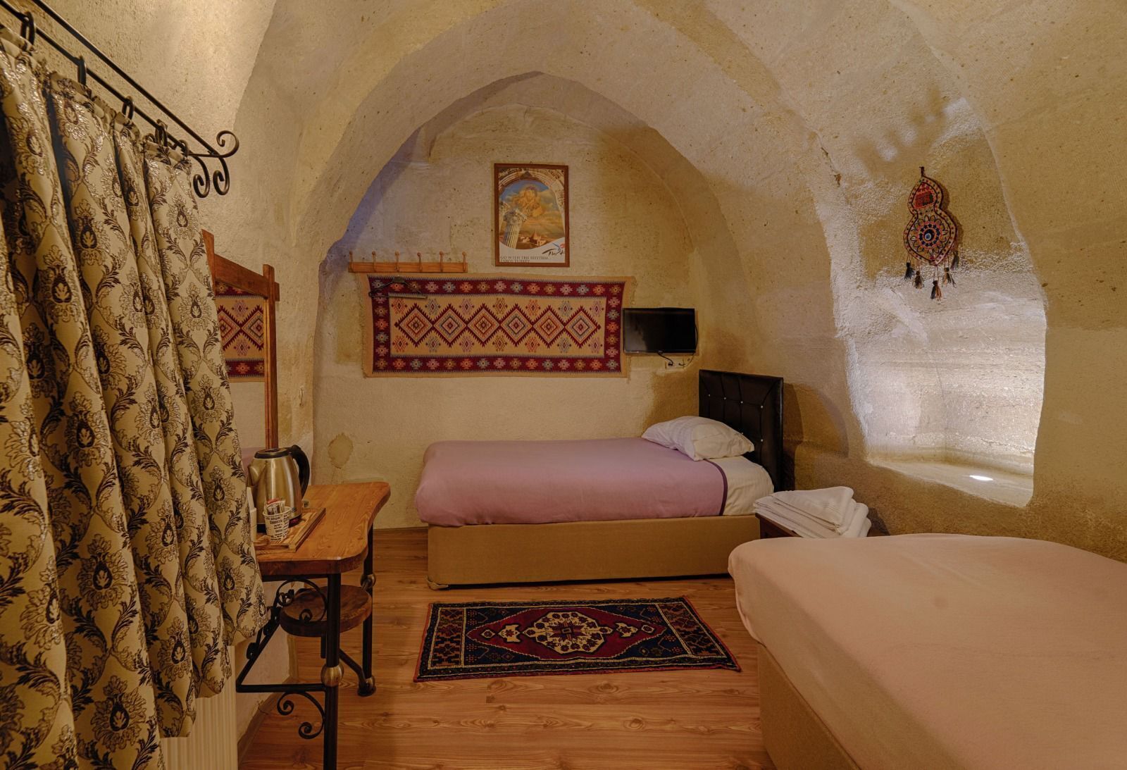 Une pièce chaleureuse et rustique avec un plafond voûté, un lit recouvert d’une courtepointe rose, un bureau en bois avec une chaise, une petite table avec une théière et un tapis à motifs.