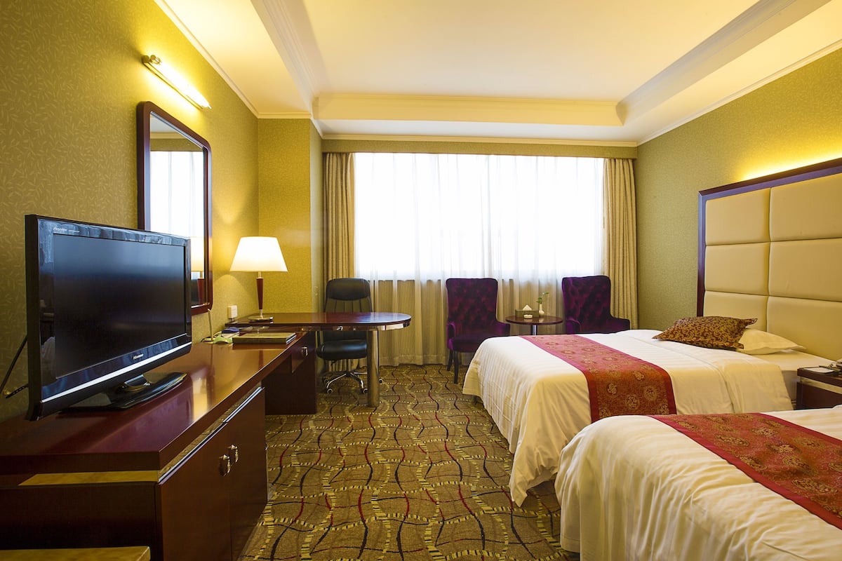Quarto de hotel com duas camas, uma escrivaninha com televisão, uma cadeira e uma janela com cortinas.