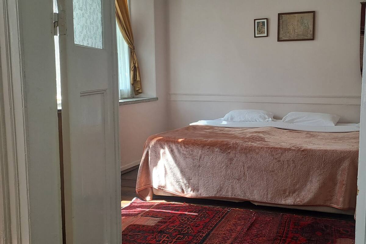 Ein Schlafzimmer mit einem Bett, einem Fenster mit Vorhängen und zwei gerahmten Bildern an der Wand.