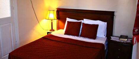 Una cama bien hecha con una manta roja, una lámpara de noche y una mesita de madera con un libro.