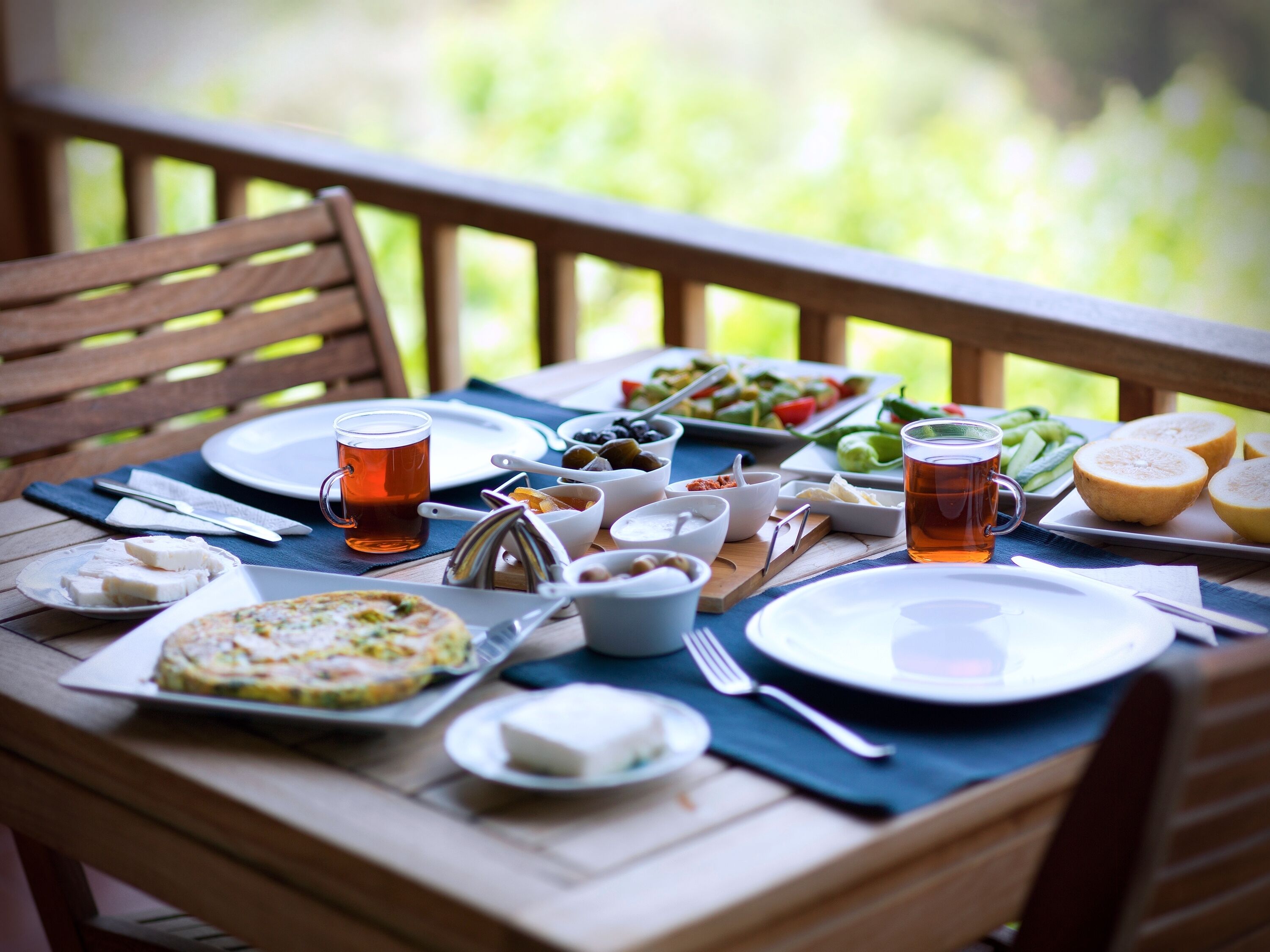 Uma mesa de madeira posta com diversos pratos, incluindo uma salada, queijo e uma fatia de pão, em um pátio externo.