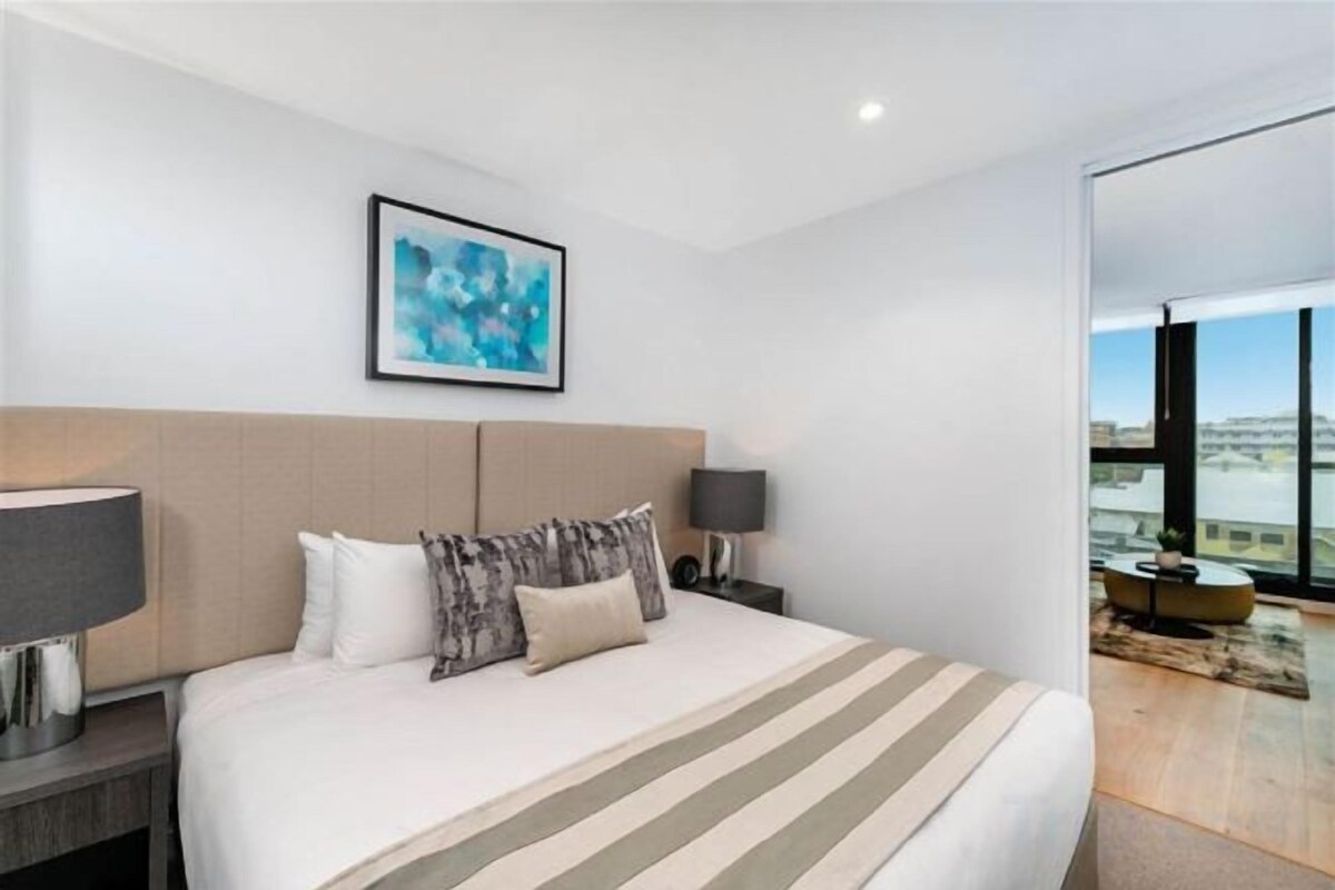 A modern hotel room with a large bed, a headboard, a nightstand with a lamp, and a view through a glass door.