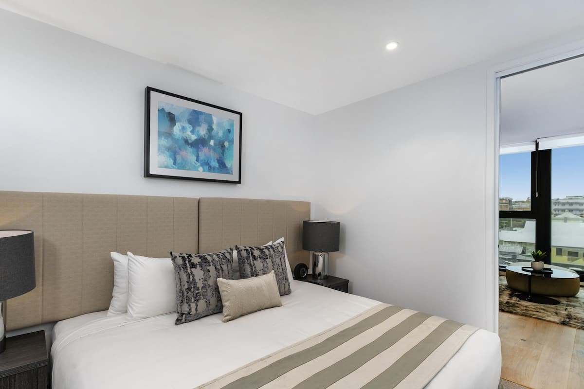 A modern hotel room with a large bed, a nightstand, a lamp, a framed abstract painting, and a view of the city through a large window.