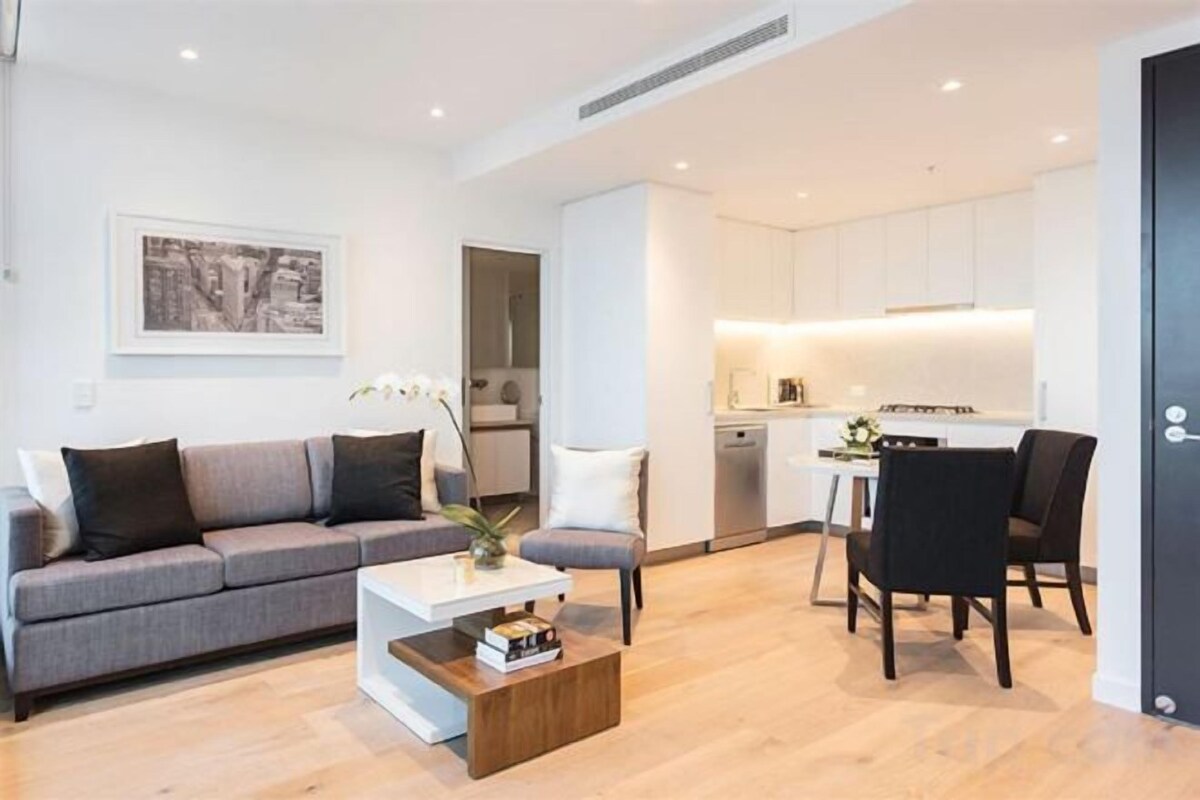 A modern living room with a sofa, coffee table, and chairs. There is a kitchen area with white cabinets and a dining table.