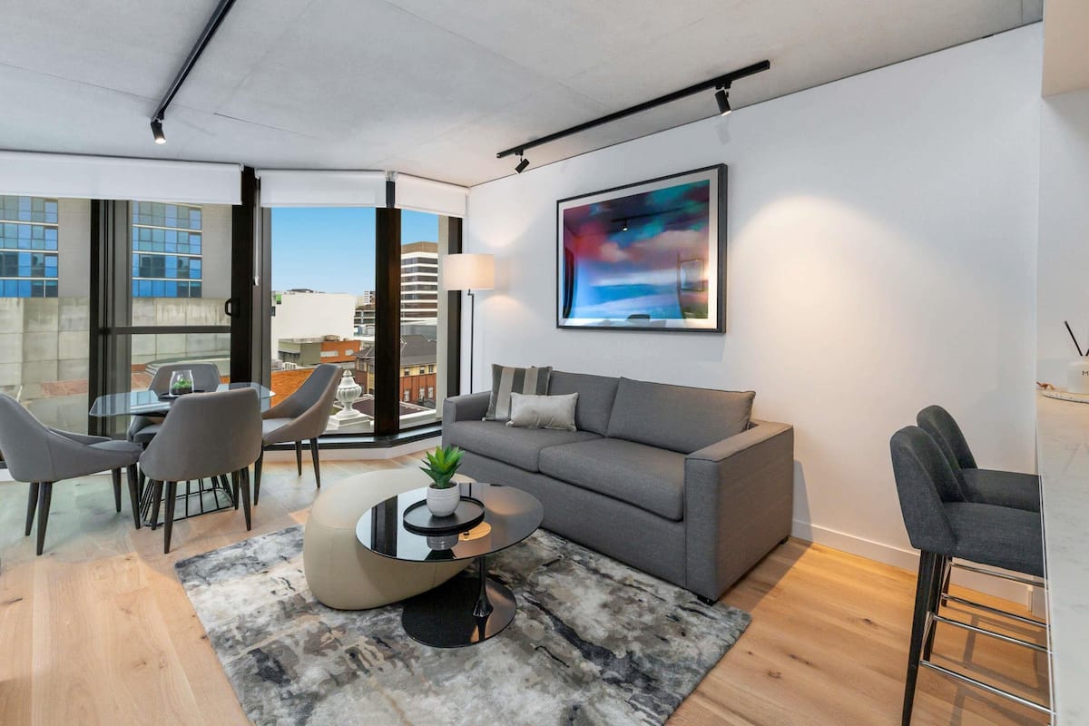 A modern living room with a grey sofa, a round coffee table, and a view of the city through large windows.