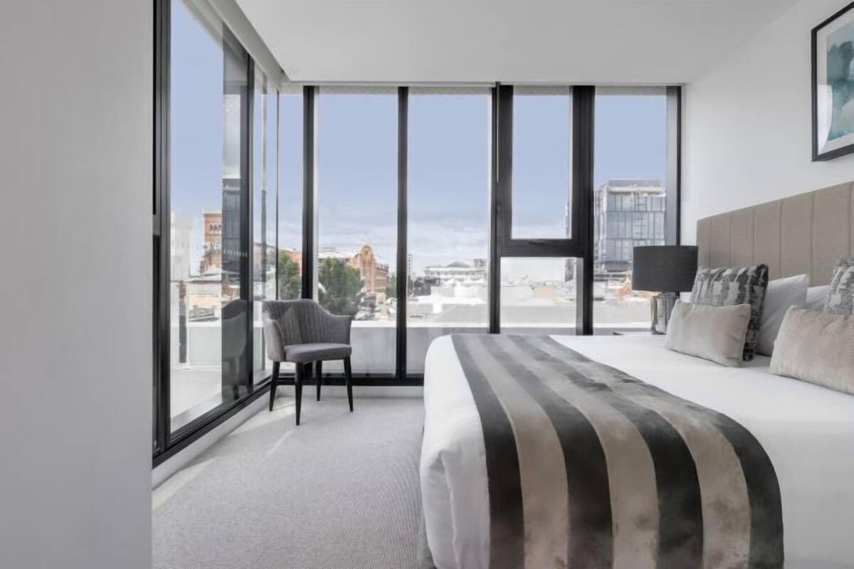 A modern hotel room with a large bed, a chair, a desk, and a view of the city through floor-to-ceiling windows.