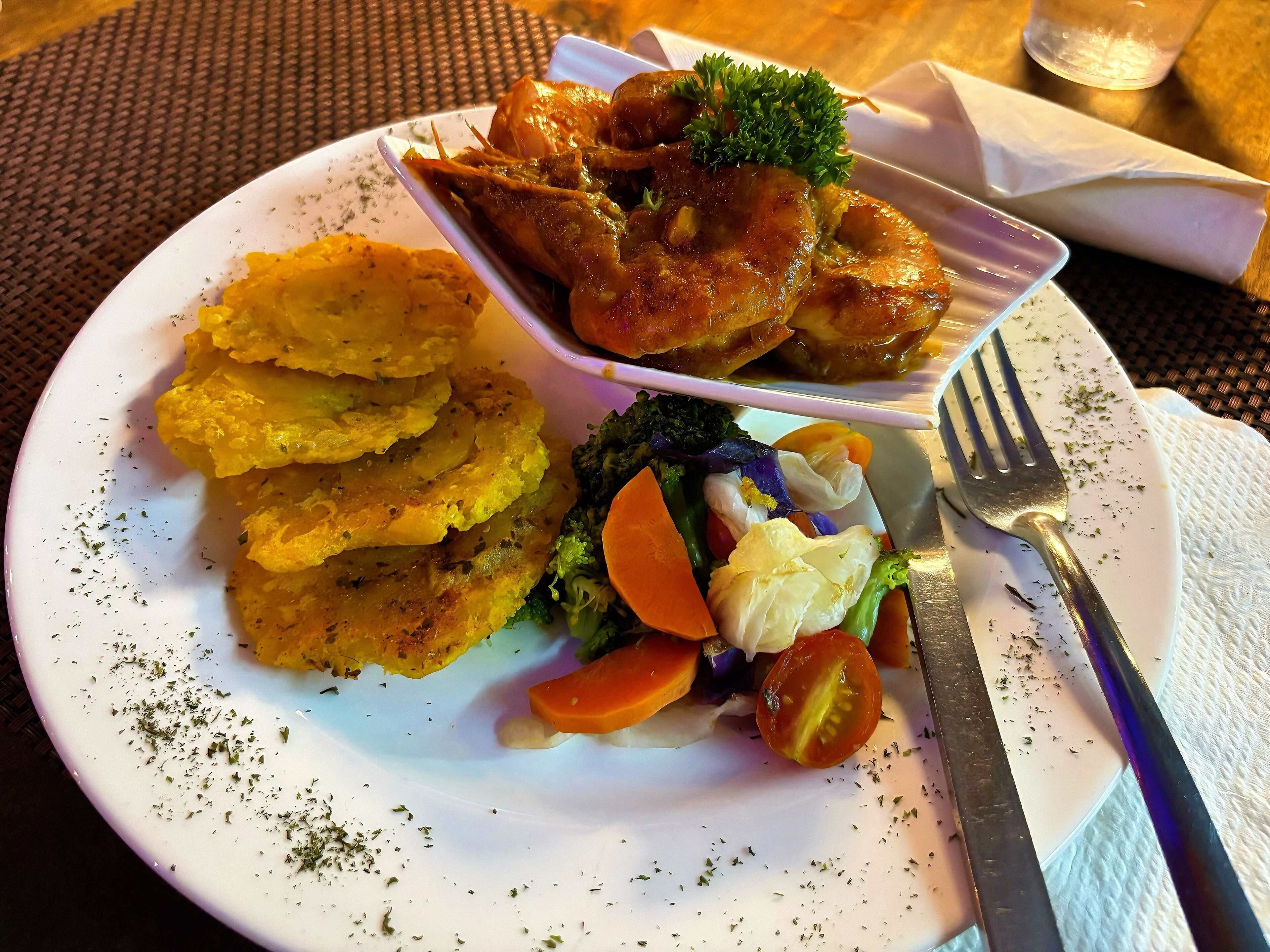 Een bord eten met diverse ingrediënten, waaronder gebakken plantains, groenten en een stuk vlees, met een vork en servetje op een tafel.