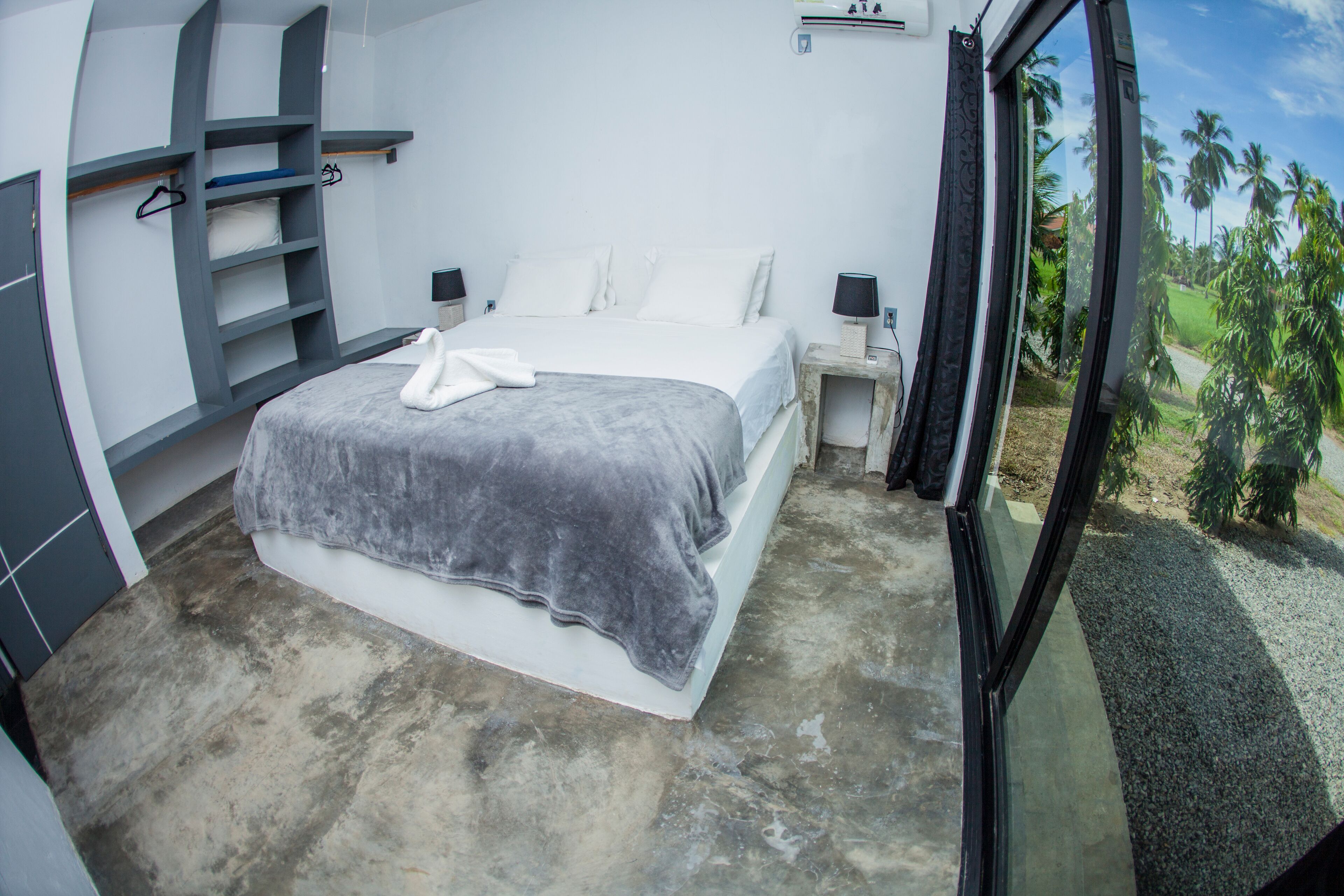 Ein modernes Schlafzimmer mit einem großen Bett, Einbauschränken, Nachttischen und Blick auf Palmen durch ein Fenster.