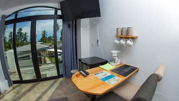 Ein Hotelzimmer mit einem Holztisch, einem an der Wand befestigten Fernseher, einer Kaffeemaschine und Blick auf Palmen durch die Glastür.
