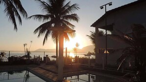Zona de piscina con vistas al atardecer, palmeras y un edificio con balcón.