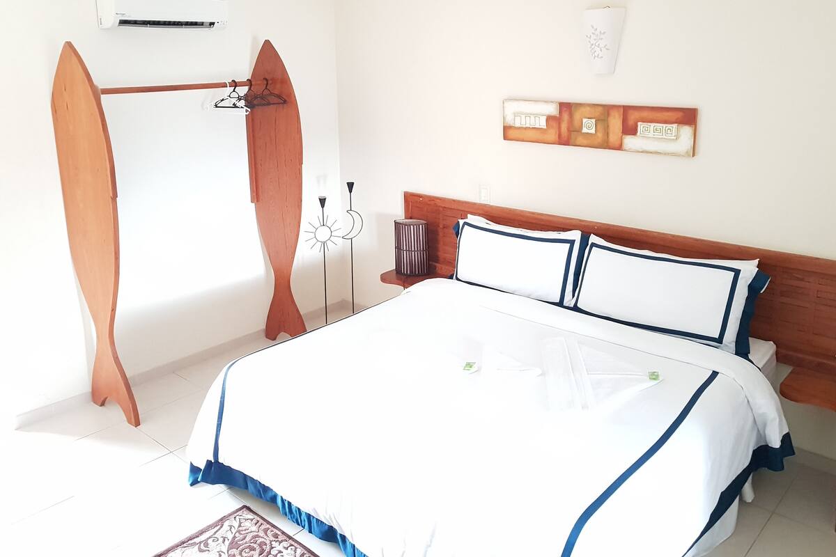 Quarto com cama, cabeceira de madeira, ar condicionado fixado na parede e uma pequena prateleira com um objeto decorativo.