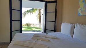 Habitación de hotel con cama, ropa de cama blanca, vista a una palmera y follaje verde afuera, y un cuadro en la pared.