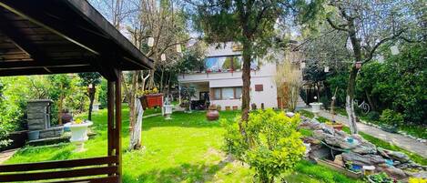 Un jardín con un banco de madera, una estructura de piedra y un edificio blanco con balcón.
