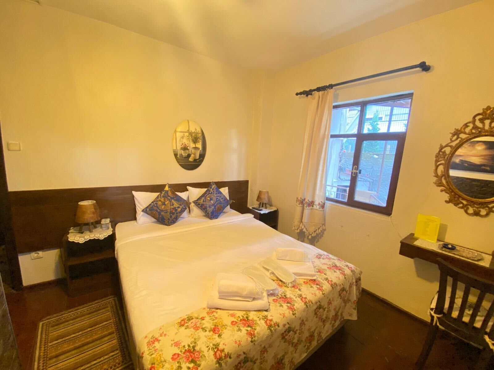 Un lit bien fait, avec une literie à motifs floraux, une table de chevet avec une lampe, une fenêtre avec des rideaux et un miroir accroché au mur.