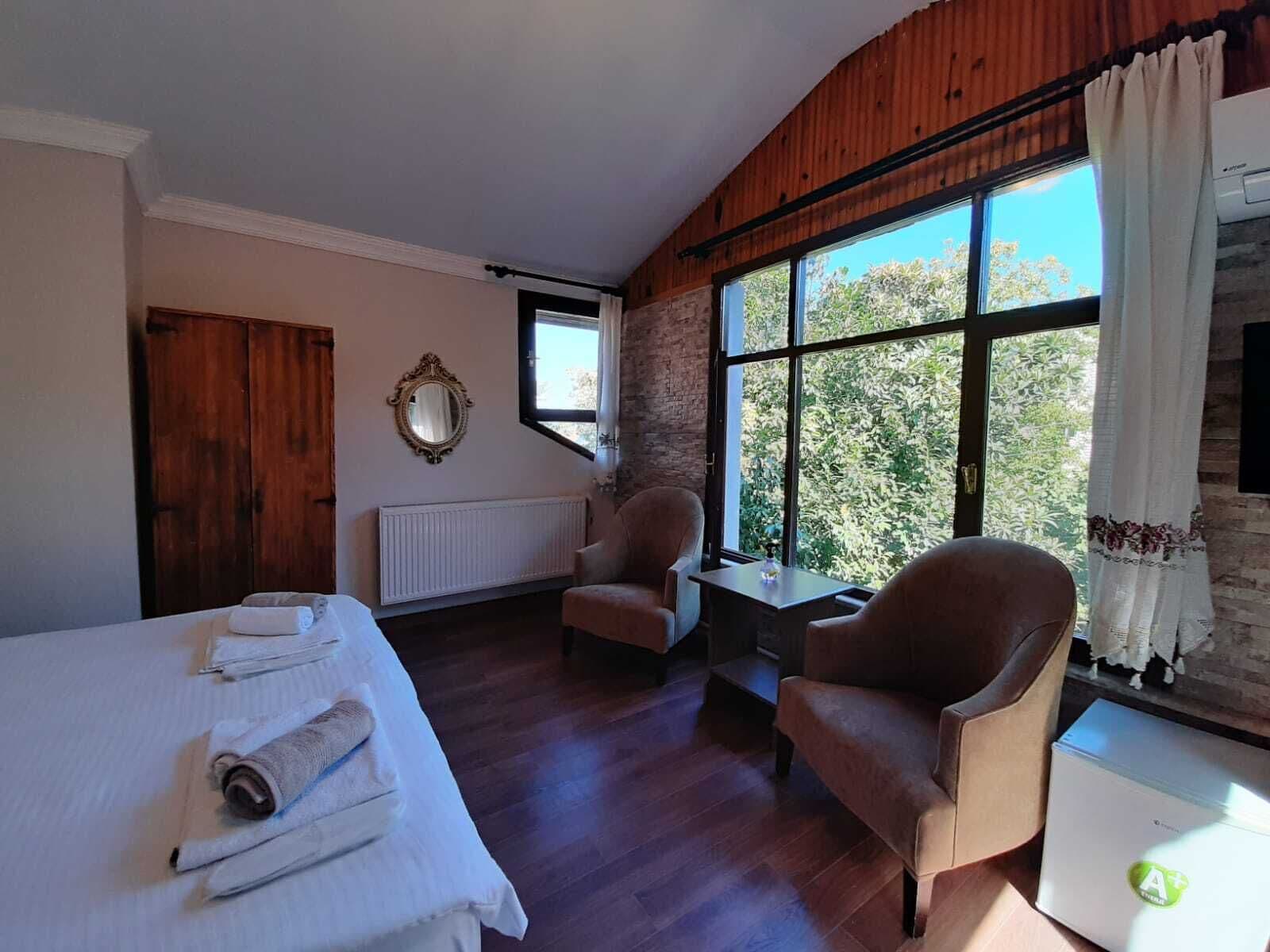 A hotel room with a bed, two armchairs, a small table, a radiator, a window with curtains, and a mirror on the wall.