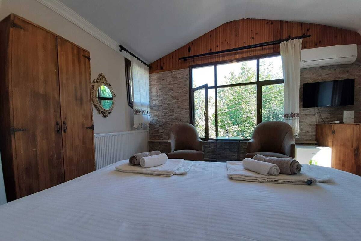 A bedroom with a large bed, two armchairs, a stone wall, a wooden ceiling, and a television.