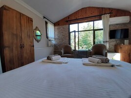 A bedroom with a large bed, two armchairs, a stone wall, a wooden ceiling, and a television.