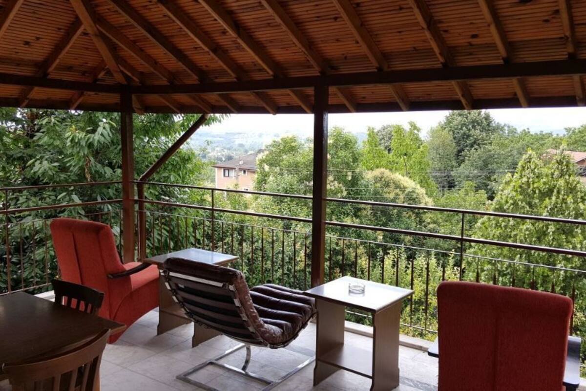 A covered balcony with a table, two chairs, and a hanging chair overlooking a lush garden and distant buildings.