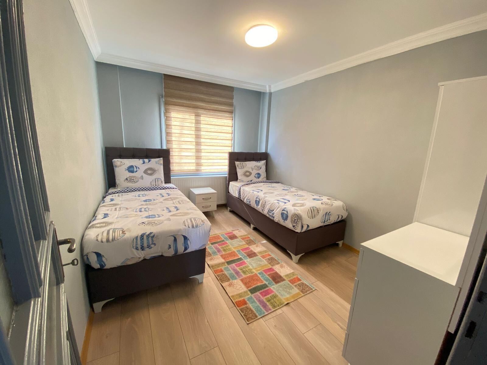 Un dormitorio con dos camas, una ventana con persianas, una alfombra colorida y un armario blanco.