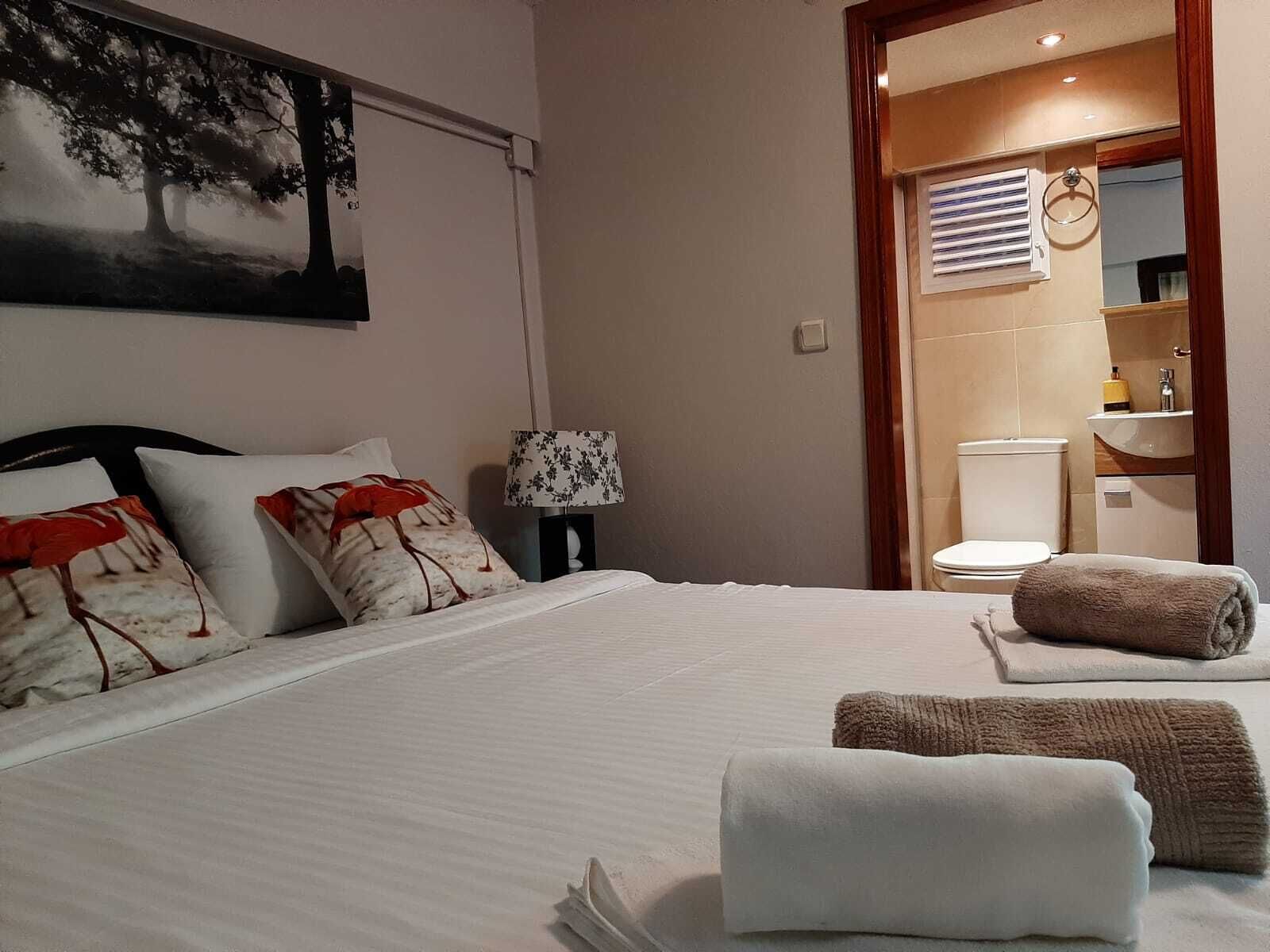 A neatly made bed with white linens and decorative pillows, a bedside lamp, and a view into a bathroom with a toilet and sink.