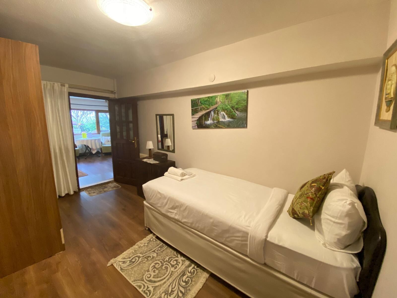 A bedroom with a bed, a wooden door, a mirror, a painting, and a view of a dining area.