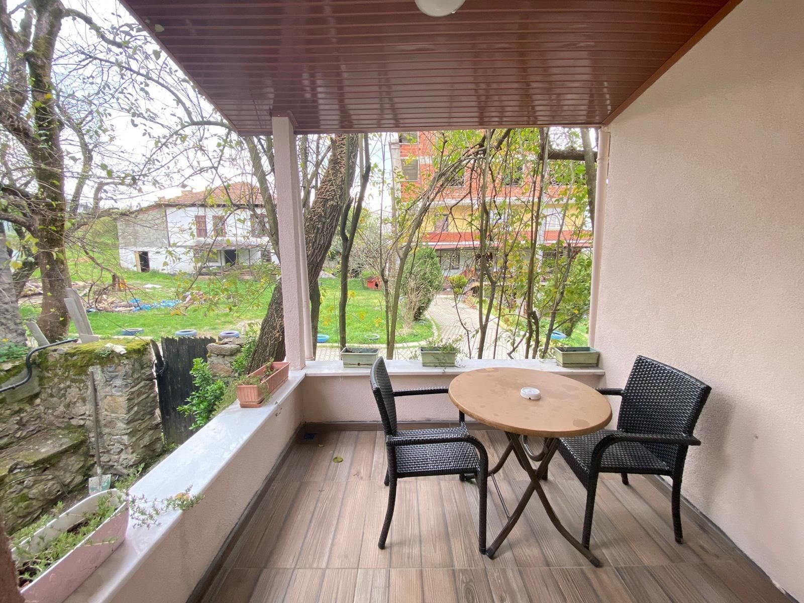 Une terrasse couverte avec une table ronde et deux chaises, donnant sur un jardin avec des arbres et un bâtiment en arrière-plan.
