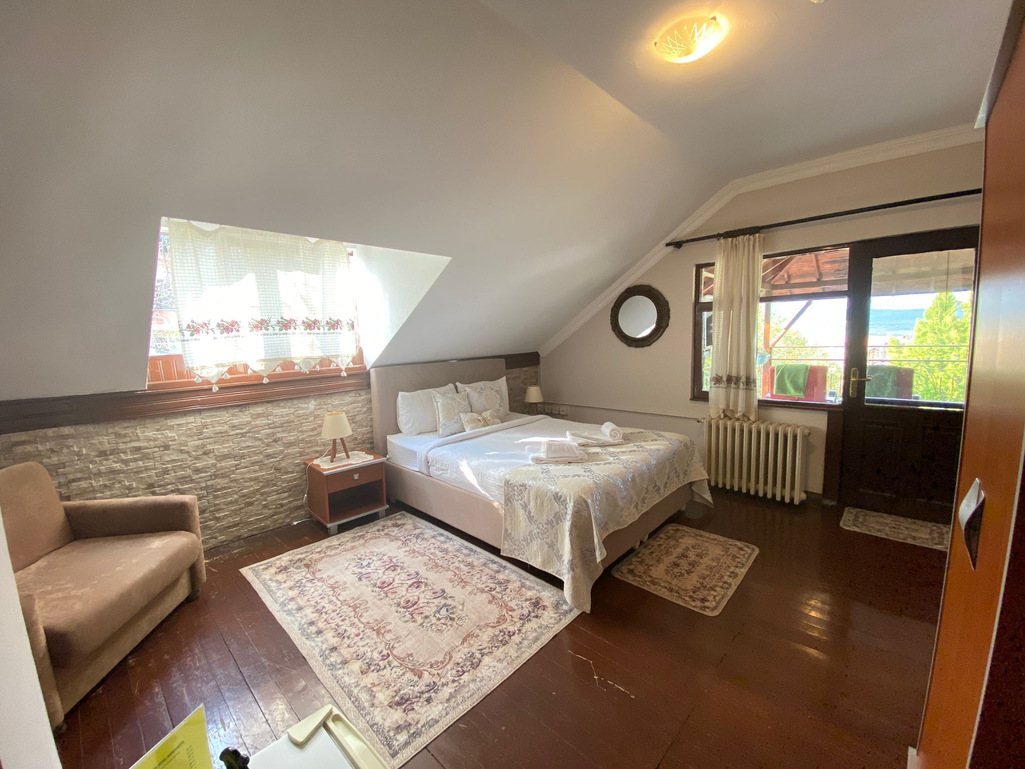 A bedroom with a sloped ceiling, a bed with white linens, a nightstand, a chair, a large window with curtains, and a view of greenery outside.