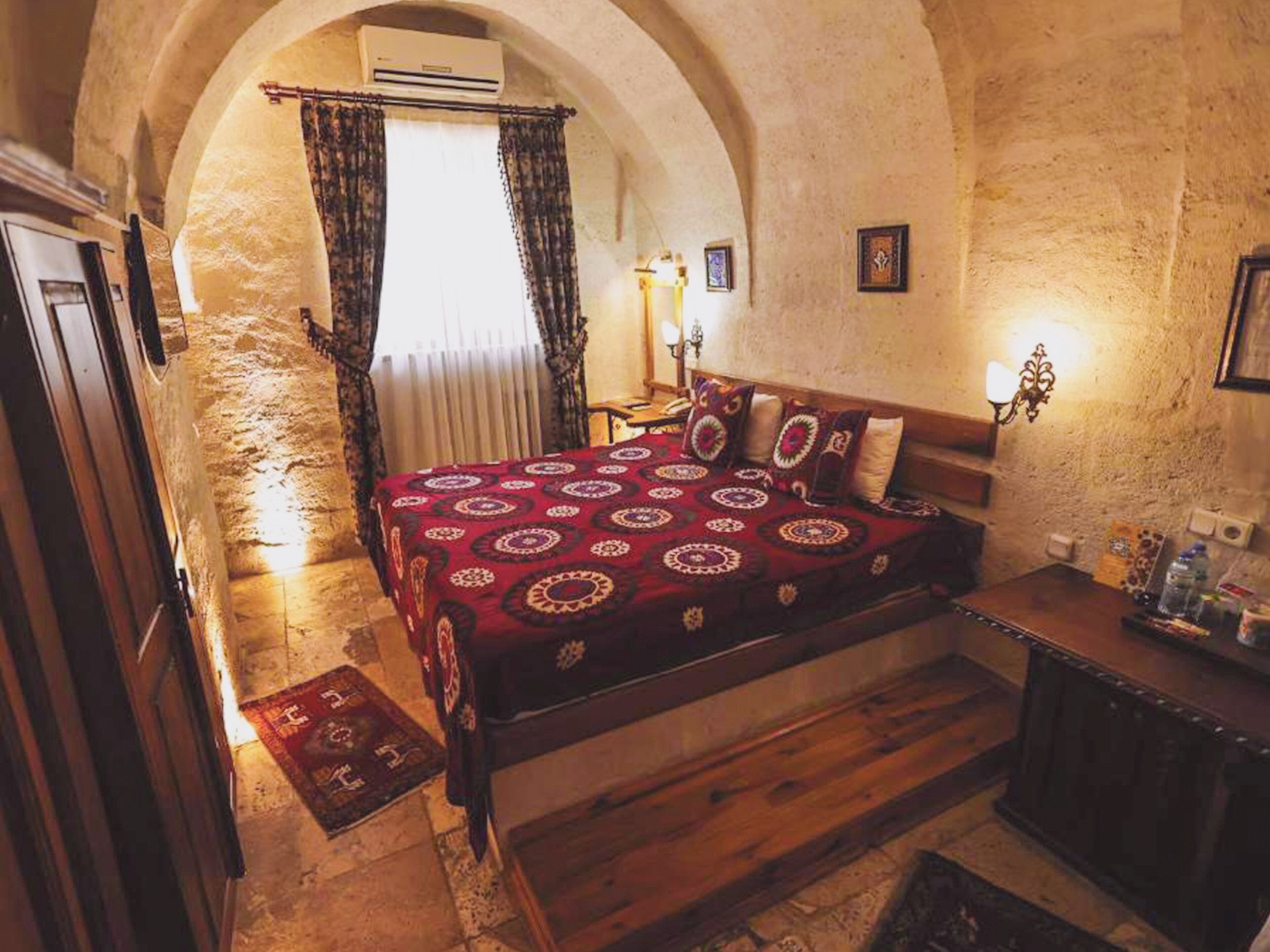 A room with a bed, a wooden dresser, a nightstand, and a small table. There is a window with curtains, a wall-mounted air conditioner, and a framed picture on the wall.
