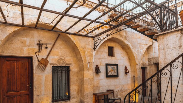 An arched courtyard with a wooden door, a window with iron bars, a small table with a chair, and a staircase with iron railings. - Hanzade Cappadocia  (Nevsehir)