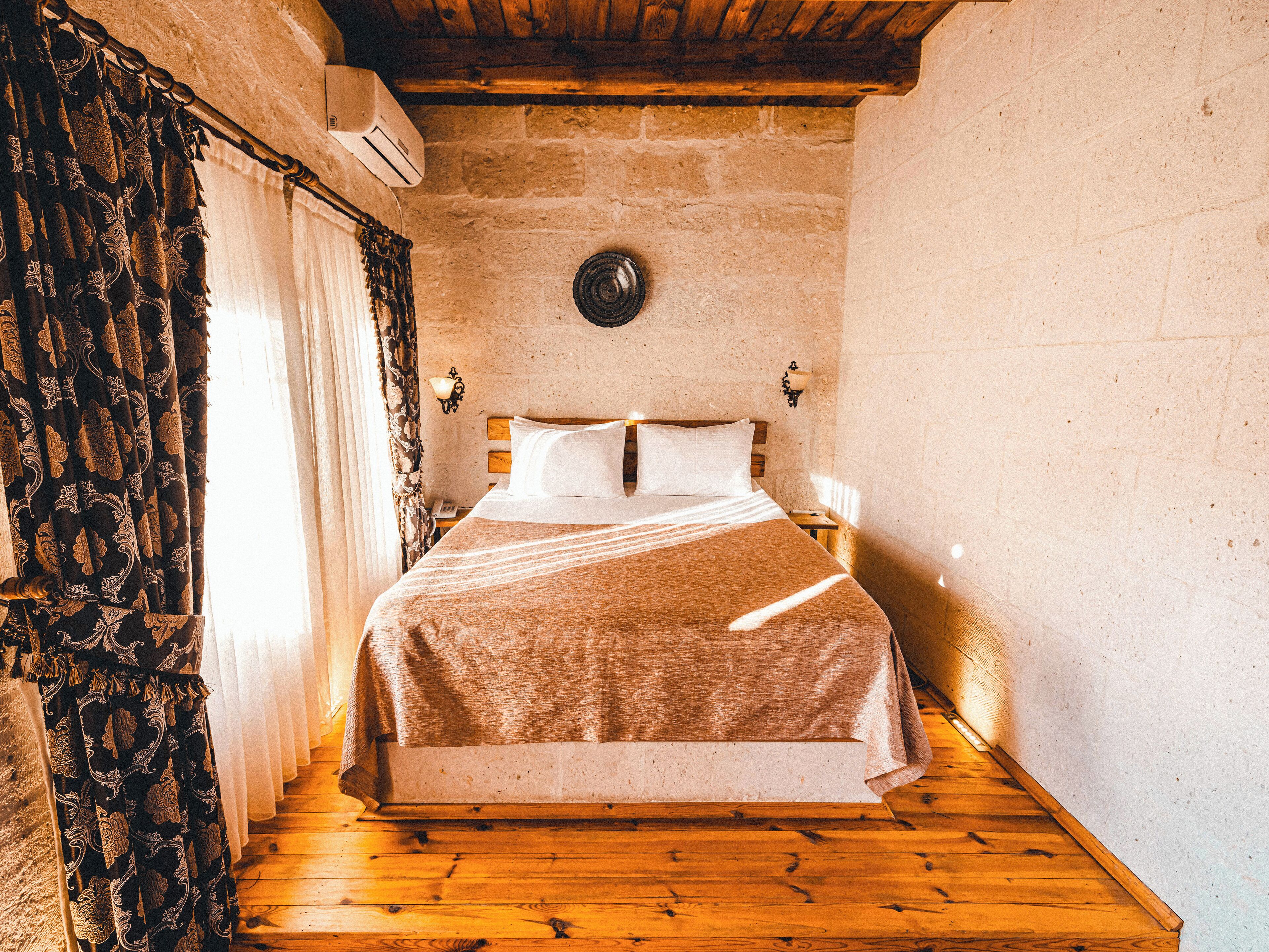 A room with a bed, wooden flooring, stone walls, and a window with curtains.